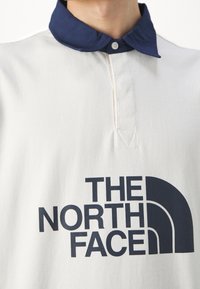 Bílá polo tričko s námořnickou modrou límečkem na knoflíky a logem "THE NORTH FACE" v černé barvě potisknutým přes hrudník.