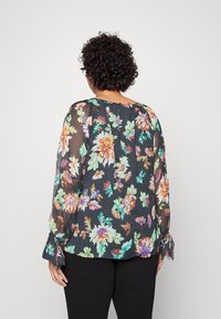 Blomsterbluse med en mørk baggrund, der har levende orange, grønne og lilla blomster. Gennemsigtigt stof, lange ærmer og en løs pasform.