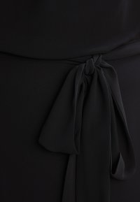 Černý textilní top s detailem zavázaného pasu. Hladká textura, zdobená uzlem a dvěma volnými konci, které vytvářejí vrstvený efekt.
