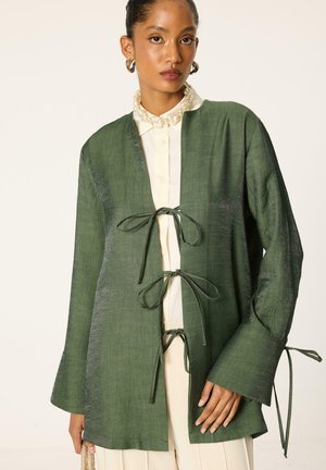 Donna che indossa una giacca aperta verde scuro con trama, legata con tre fiocchi sopra una camicetta color crema e una gonna plissettata, accessoriata con una collana di perle.