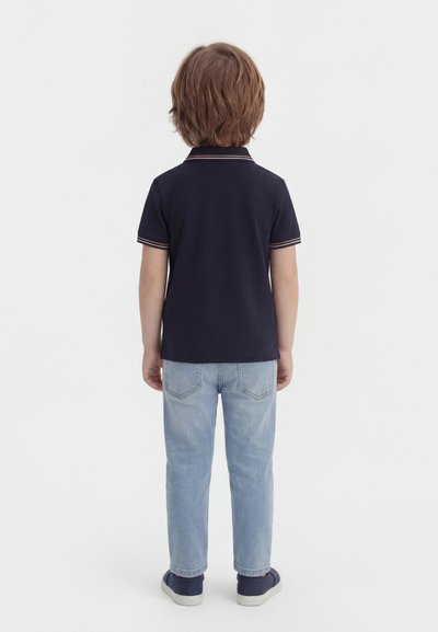 Παιδί στέκεται γυρισμένο ανάποδα, φοράει μπλε πουκάμισο πόλο, ανοιχτόχρωμο τζιν και σκούρα αθλητικά παπούτσια σε λευκό φόντο.