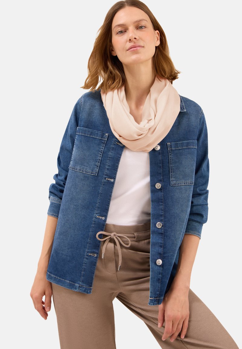 Chemise en denim bleu à boutons, comprenant deux poches appliquées et des boutons argentés. Portée par-dessus un haut blanc et assortie à un pantalon beige à cordon de serrage et à une écharpe légère.