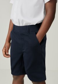Marineblauwe shorts van een textuurstof, met zijzakken en een platte voorkant. G gedragen met een witte overhemd met kraag.