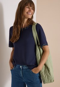 Smiling woman with long brown hair wearing a navy shirt with leopard print trim and blue jeans, carrying a quilted light green tote bag.