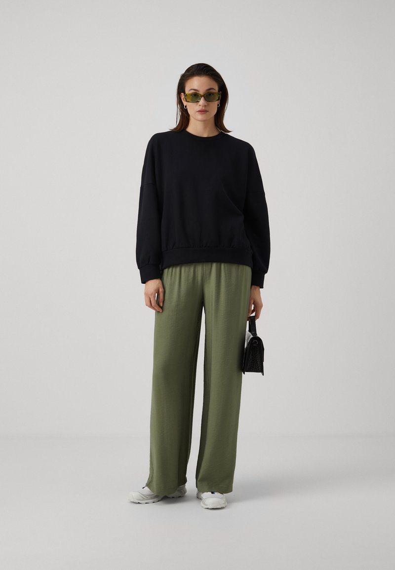 Felpa nera oversize con orlo a coste, abbinata a pantaloni larghi verde oliva. Il modello indossa occhiali da sole verdi e tiene una piccola borsa nera.