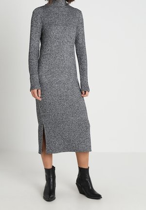 Kobieta ubrana w szarą prążkowaną sukienkę midi z rozcięciem z boku oraz czarne botki, stojąca na jednolitym jasnym tle.