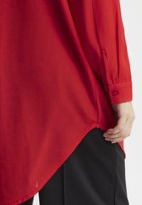 Blusa rossa in tessuto morbido, con maniche lunghe e polsini con bottone, e orlo arrotondato, che copre parzialmente i pantaloni neri.