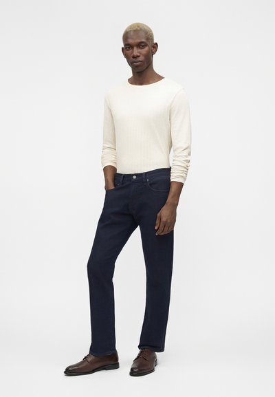 Άνδρας με κοντά ξανθά μαλλιά που φοράει μια κρεμ μακρυμάνικη μπλούζα, σκούρα μπλε τζιν και καφέ δερμάτινα παπούτσια, στέκεται με το ένα χέρι στην τσέπη.