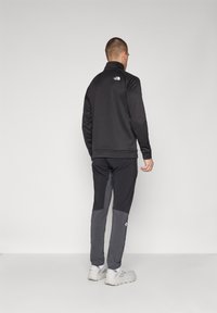 Crna fleece jakna s patentnim zatvaračem i visokim ovratnikom, s logom na leđima, u kombinaciji sa sivim hlačama koje imaju dizajn u boji.