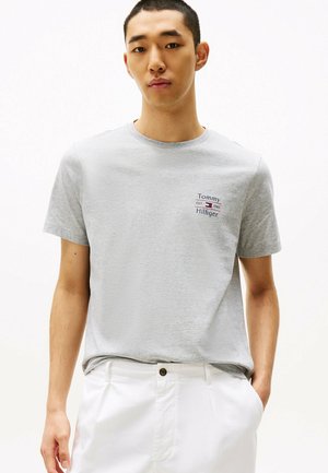 T-shirt en coton gris avec un petit logo Tommy Hilfiger brodé. Manches courtes et encolure ronde. Porté avec un pantalon blanc.