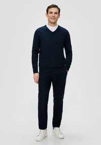 Dunkelblauer V-Ausschnitt-Pullover über einem weißen T-Shirt, kombiniert mit dunkelblauen Hosen und weißen Sneakers. Das Material wirkt weich und strukturiert.