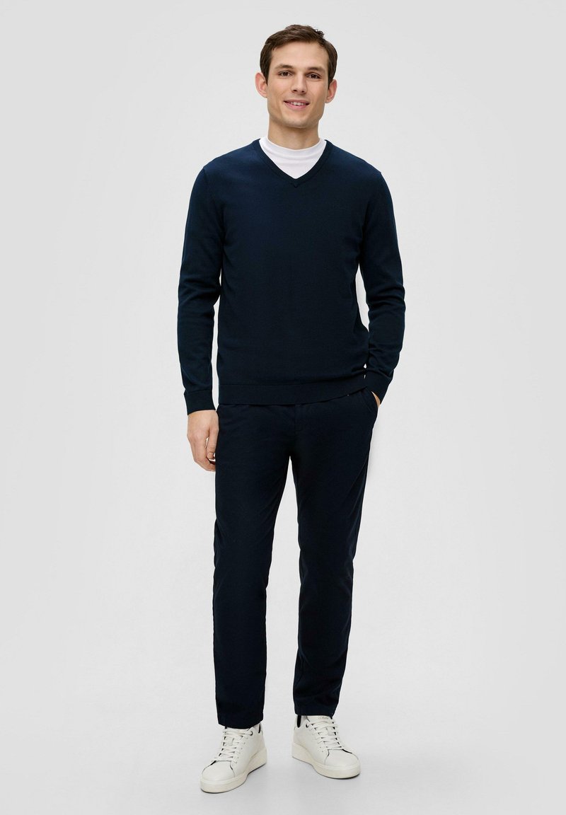 Dunkelblauer V-Ausschnitt-Pullover über einem weißen T-Shirt, kombiniert mit dunkelblauen Hosen und weißen Sneakers. Das Material wirkt weich und strukturiert.