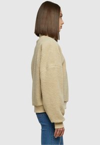 Veste en polaire beige avec un col montant, coupe surdimensionnée et poignets côtelés. Le modèle l'a associée à un jean ajusté, présentée de profil.