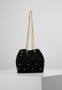 Borsa a secchiello in suede nera con tracolle in catena dorata, caratterizzata da borchie quadrate sparse sulla superficie. Texture morbida e forma arrotondata.