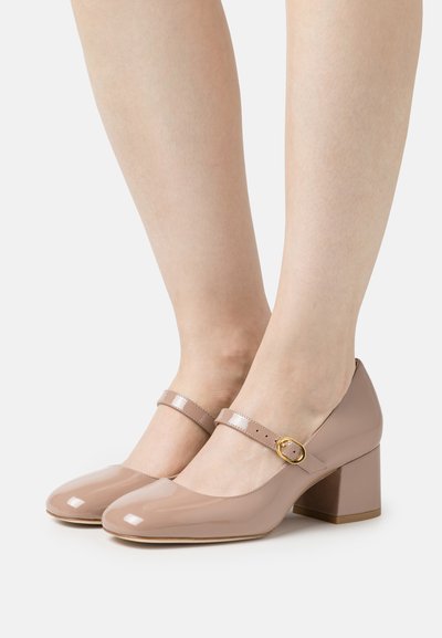 Bottines Mary Jane en cuir verni beige avec un bout arrondi, un talon bloc et une boucle décorative sur une sangle autour de la cheville.