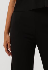 Pantaloni neri su misura con una texture liscia e una vestibilità tailored, caratterizzati da un vita media e un dettaglio con spacco laterale. Mano sulla vita.