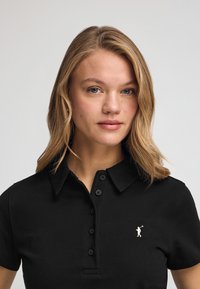 Camisa polo negra hecha de tela texturizada con cuello, con una tapeta con botones y una pequeña figura bordada en el lado izquierdo del pecho.