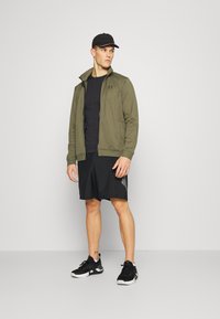 Olivgrüne Zip-Jacke, schwarzes T-Shirt, schwarze Shorts mit seitlichem Muster und schwarze Turnschuhe. Das Model steht vor einem neutralen Hintergrund.