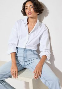 Camisa blanca de botones con mangas arremangadas, combinada con jeans de cintura alta de color azul claro. Modelo sentado sobre una superficie de concreto texturizada.