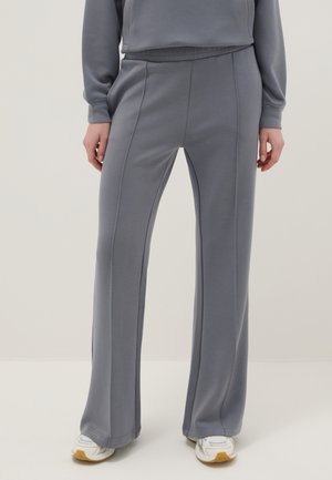 Persona con pantalones anchos grises y top a juego con zapatillas blancas, de pie frente a un fondo claro y liso.