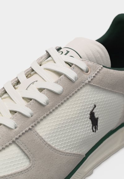 Gros plan sur une chaussure de sport beige et blanche avec des lacets blancs, un panneau latéral texturé et un logo Polo noir sur le côté.