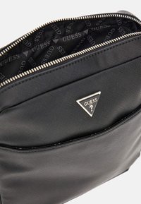 Bolso de mano sintético negro con un acabado texturizado, que presenta una cremallera plateada y un logotipo triangular en el bolsillo frontal. Interior forrado con un diseño de logotipos.