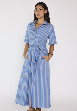 Femme debout, les mains dans les poches, portant une robe midi à rayures verticales bleu et blanc, à manches courtes et ceinturée à la taille.