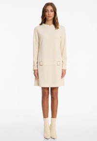 Beige langärmliges Kleid mit einem schlichten Schnitt, das über einen kleinen Ausschnitt und zwei quadratische Akzente an der Taille verfügt. Sanfte Textur, knielang.