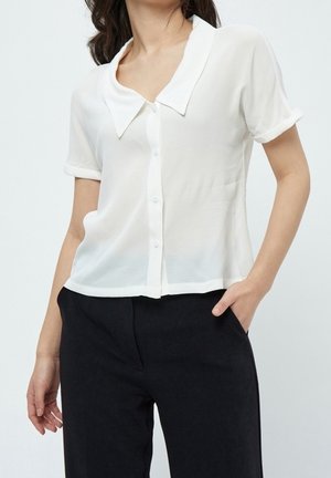 Kobieta w białej, krótkorękawowej bluzce zapinanej na guziki z szerokim kołnierzem i czarnych spodniach, stojąca z jedną ręką w kieszeni.