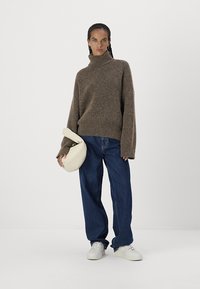 Maglione a collo alto marrone in lana con maniche lunghe, abbinato a jeans a gamba larga blu e sneakers bianche, mentre porta una borsa color crema.