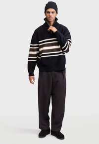 Pulover tricotat negru cu dungi orizontale bej și maro, guler cu jumătate de fermoar, asociat cu pantaloni închiși la culoare și o căciulă asortată.