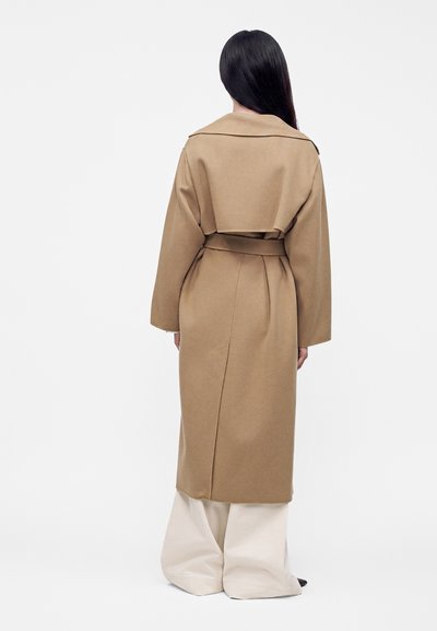Femme aux longs cheveux foncés portant un trench-coat camel ceinturé et un large pantalon blanc, debout de dos sur un fond uni.