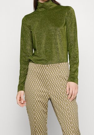 Žena v zeleném třpytivém roláku s dlouhým rukávem a béžových kalhotách s bílým a zeleným diamantovým vzorem.