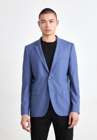 Sacou bleu deschis cu rever în formă de v, închidere cu un singur nasture, două buzunare frontale și un buzunar pe piept, purtat peste o cămașă neagră.