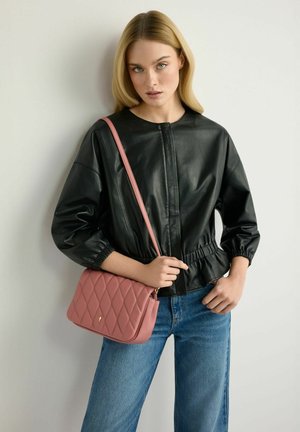 Kobieta o prostych blond włosach, ubrana w czarną skórzaną kurtkę i niebieskie dżinsy, trzymająca pikowaną różową torebkę na ramię.