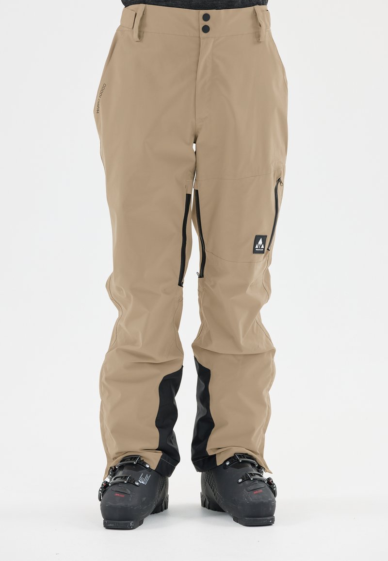 Pantalons de ski beige avec fermetures éclair et accents noirs, présentant une coupe ample et un matériau imperméable. Comprend un détail de logo sur le côté.