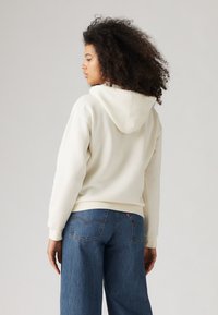 Kvinna med lockigt hår som bär en krämfärgad hoodie och blå jeans, stående vänd åt sidan mot en ljus enhetlig bakgrund.