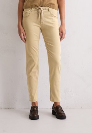 Vrouw in beige slim-fit broek met een trekkoord in de taille en een paar bruine leren instappers, staand tegen een witte, textuurrijke muur.