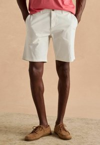 Heledad hall tanžaadiga puuvillased lühikesed püksid küljetaskutega, sirge lõike ja ülevalt keeratud äärega, kombineerituna pruunide sametmaterjalist kingadega.