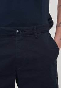 Pantaloni in cotone blu navy con chiusura con bottone, tasche laterali e una texture liscia. Presentano linee pulite e una vestibilità sartoriale.