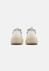 Weiße und beige Sneakers mit einer kräftigen Sohle, die eine strukturierte Rückseite mit geprägtem Branding und subtilen grauen Akzenten in der Nähe der Zunge aufweisen.