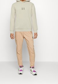 Felpa grigia chiaro con tasca a marsupio e cordino, abbinata a pantaloni cargo pesca con tasche laterali. Sneakers sportive bianche con accenti viola.