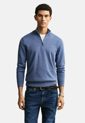 Mann steht und trägt einen blauen Pullover mit Reißverschluss bis zur Brust über einem weißen T-Shirt, dunkle Jeans, einen geflochtenen braunen Gürtel und eine silberne Armbanduhr.