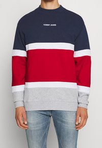 Muž v mikině s kulatým výstřihem v námořnické, bílé, červené a šedé barvě, s nápisem "TOMMY JEANS" na hrudi, spárované s světle modrými džíny.