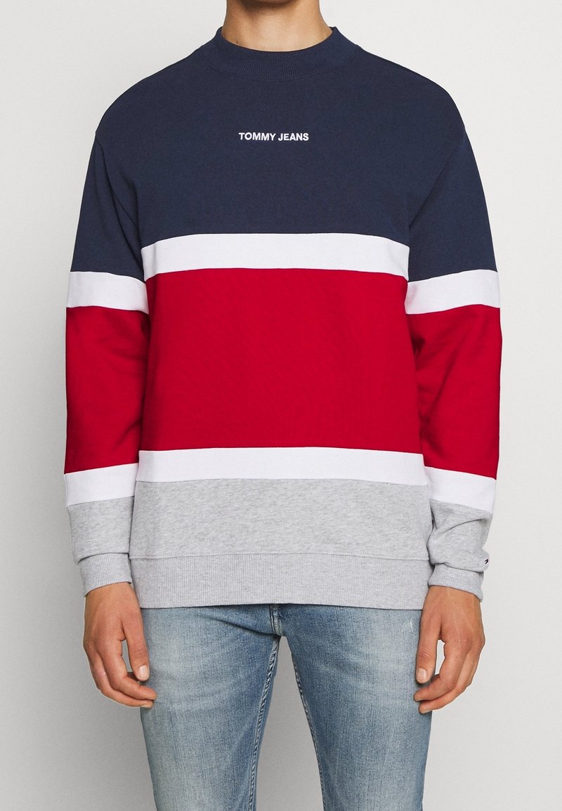 Muž v mikině s kulatým výstřihem v námořnické, bílé, červené a šedé barvě, s nápisem "TOMMY JEANS" na hrudi, spárované s světle modrými džíny.