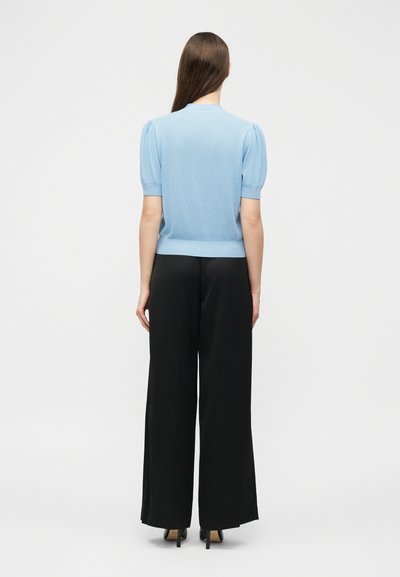 Femme debout de dos, portant un haut bleu clair à manches courtes avec des épaules bouffantes et un pantalon noir à jambes larges avec des talons.