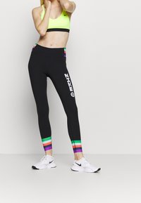 Leggings negros con rayas horizontales de colores en el dobladillo y un logo blanco de "Nike" en el costado. Combinados con un sujetador deportivo amarillo brillante.