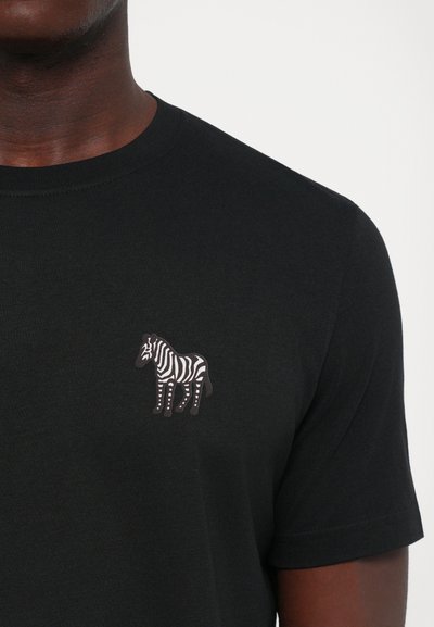 Melnā T-krekls, ko valkā persona, ar nelielu zebras grafiku, kas iespiesta kreisajā krūtīs.