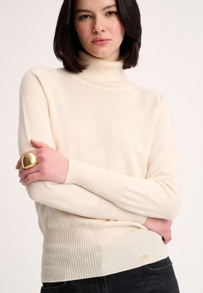 Pulover crem cu guler înalt și manșete și tiv cu textură ondulată, având o croială ajustată și o textură subtilă. Modelul poartă o brățară de aur. 