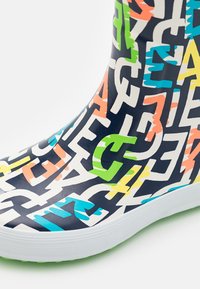 Farverig regnstøvle med store, overlappende bogstaver i blå, orange, grøn og gul på en mørk baggrund; hvid sål med grønne detaljer.